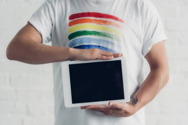 cropped shot of man in t-shirt with pride flag holding tablet with blank screen in front of white brick wall clipart