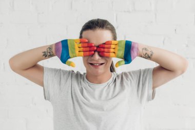 smiling transgender woman covering eyes with hands painted in colors of pride flag in front of white brick wall clipart