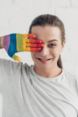 smiling transgender woman covering eye with hand painted in colors of pride flag in front of white brick wall clipart