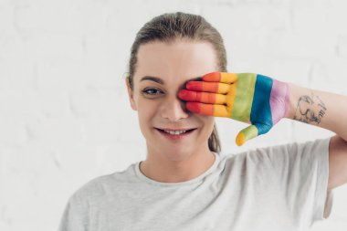 young transgender woman covering eye with hand painted in colors of pride flag in front of white brick wall clipart