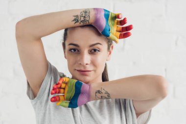 young transgender woman with hands painted in colors of pride flag in front of white brick wall clipart