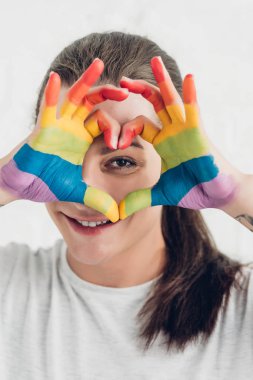young transgender woman looking at camera while making heart sign with hands in colors of pride flag in front of white brick wall clipart