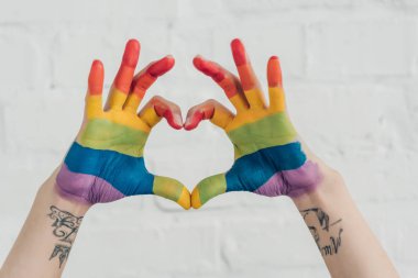 cropped shot of hands painted in colors of pride flag showing heart gesture in front of white brick wall clipart