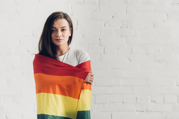 молодая трансгендерная женщина, покрытая флагом гордости и смотрящая на камеру перед стеной из белого кирпича