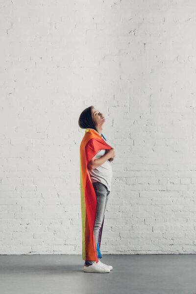 young transgender woman covering shoulders with pride flag in front of white brick wall