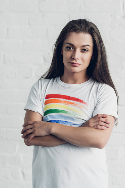 young transgender woman in white t-shirt with pride flag looking at camera with crossed arms in front of white brick wall