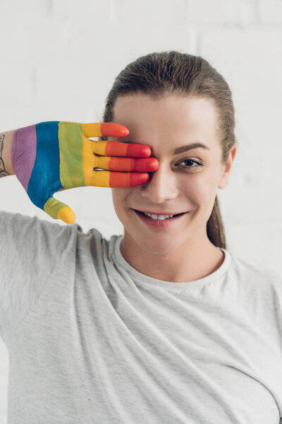 smiling transgender woman covering eye with hand painted in colors of pride flag in front of white brick wall