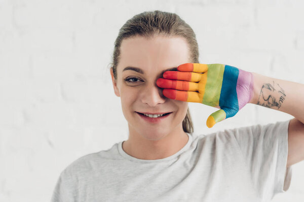 young transgender woman covering eye with hand painted in colors of pride flag in front of white brick wall