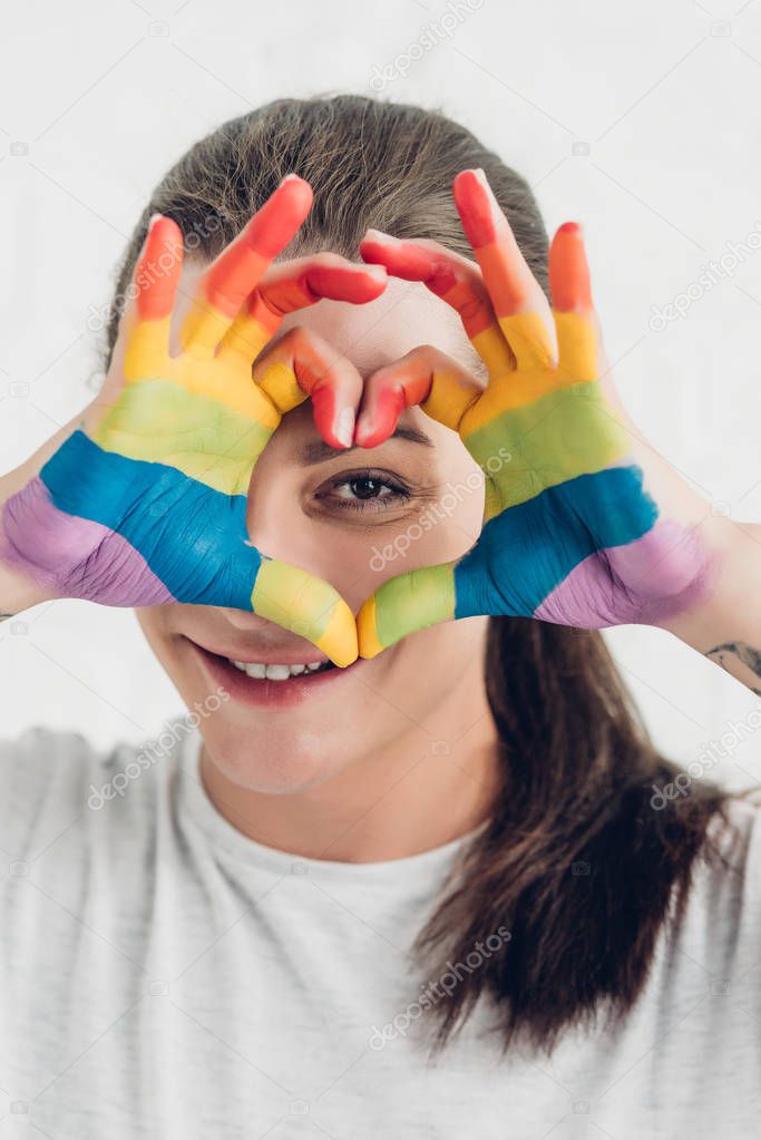 young transgender woman looking at camera while making heart sign with hands in colors of pride flag in front of white brick wall