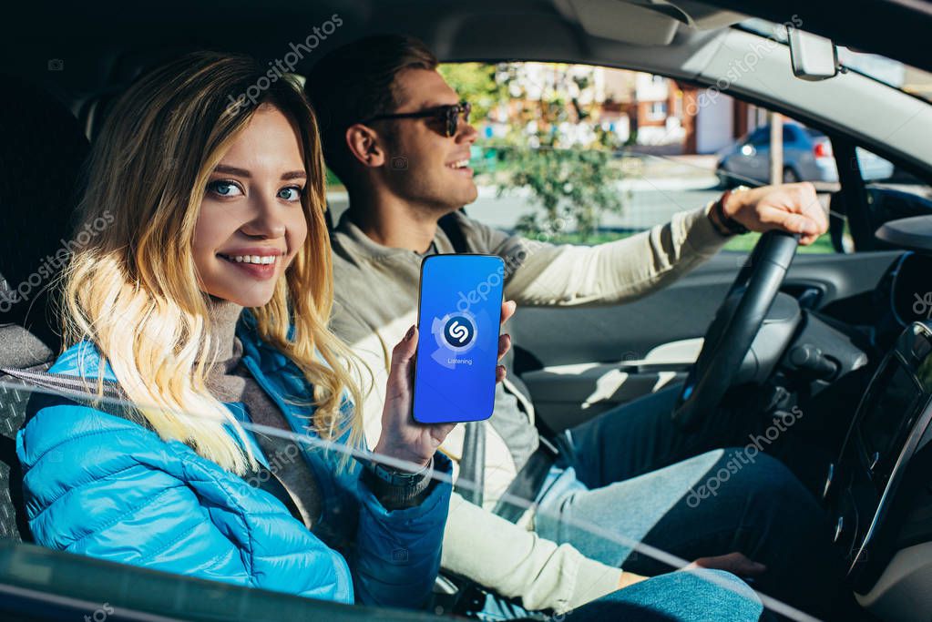 Smiling woman showing smartphone with shazam logo on screen while husband driving car