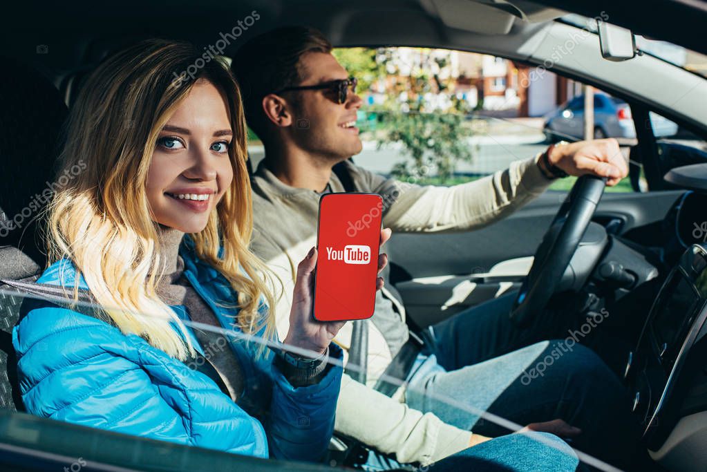Smiling woman showing smartphone with youtube logo on screen while husband driving car