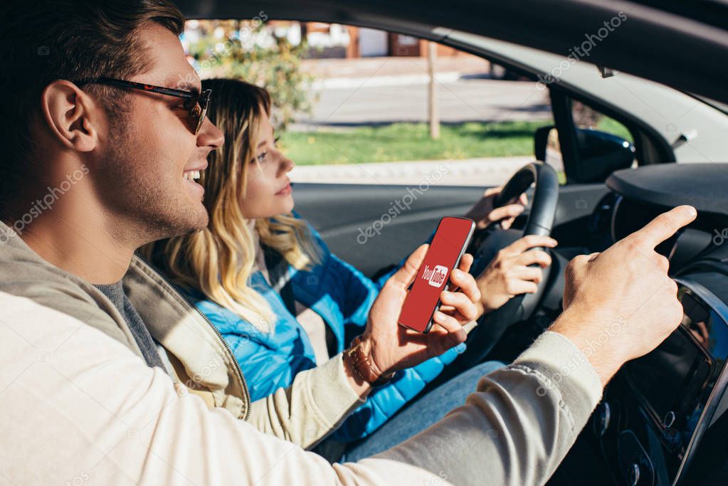 Man with smartphone with youtube logo on screen showing direction to wife  in car