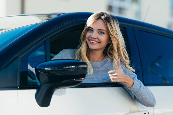 портрет белокурой улыбающейся женщины, показывающей большой палец вверх во время вождения автомобиля
