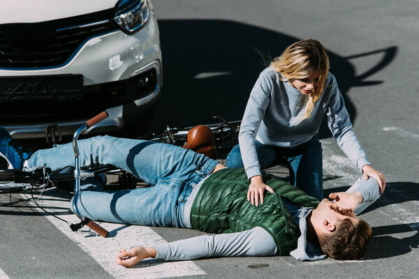 woman looking at injured young cyclist lying with bicycle on road at car accident