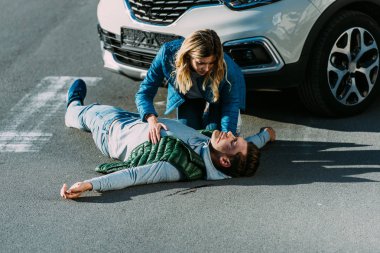 high angle view of scared young woman touching injured man lying on road after car accident clipart