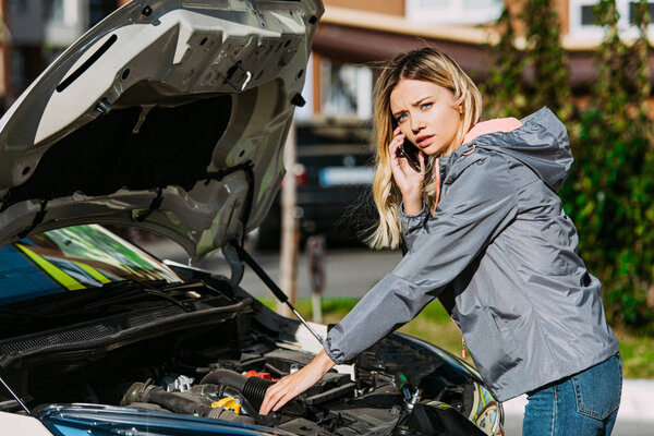 girl talking by smartphone and looking at camera while fixing broken car