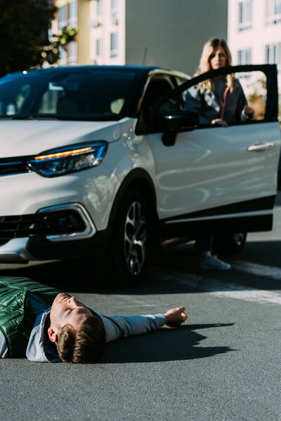 раненый молодой человек лежит на дороге, а женщина стоит позади машины после ДТП
