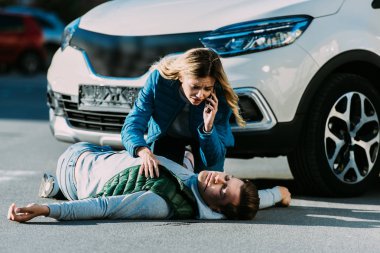 scared young woman calling emergency and looking at injured man on road after traffic accident  clipart