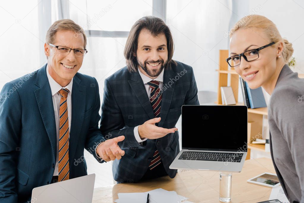 young smiling male insurance agent standing with workers and pointing at laptop in office