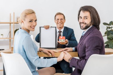 happy couple holding hands while insurance agent showing contract and small house clipart