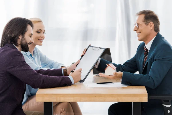 adult smiling insurance agent showing contract and small house to young couple in office