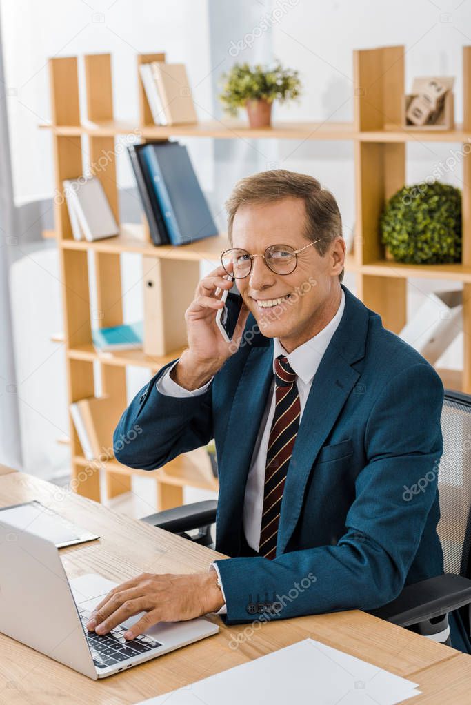 adult smiling businessman in glasses talking on smartphone and using laptop in office