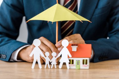 Male hands with paper cut family, house model and umbrella on wooden table, life insurance clipart
