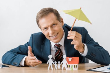 businessman holding umbrella with paper cut family and house model on wooden table clipart
