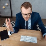 Visão de alto ângulo de homem de negócios irritado em óculos que gesticula por mãos à mesa com teclado de computador e rato no escritório