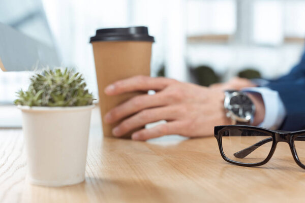 cropped image of businessman sitting with disposable coffee cup at table with potted plant and eyeglasses in modern office 