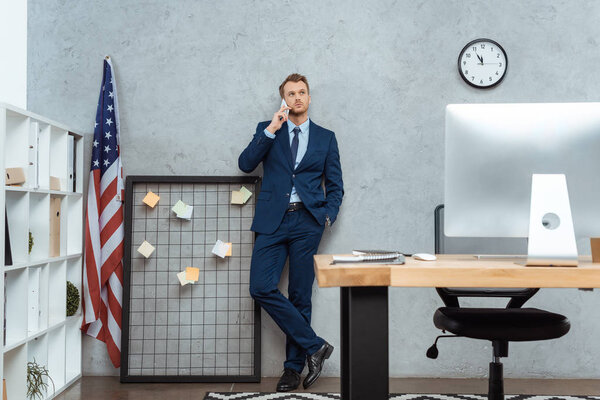 handsome businessman in suit talking on smartphone near american flag by wall in modern office 