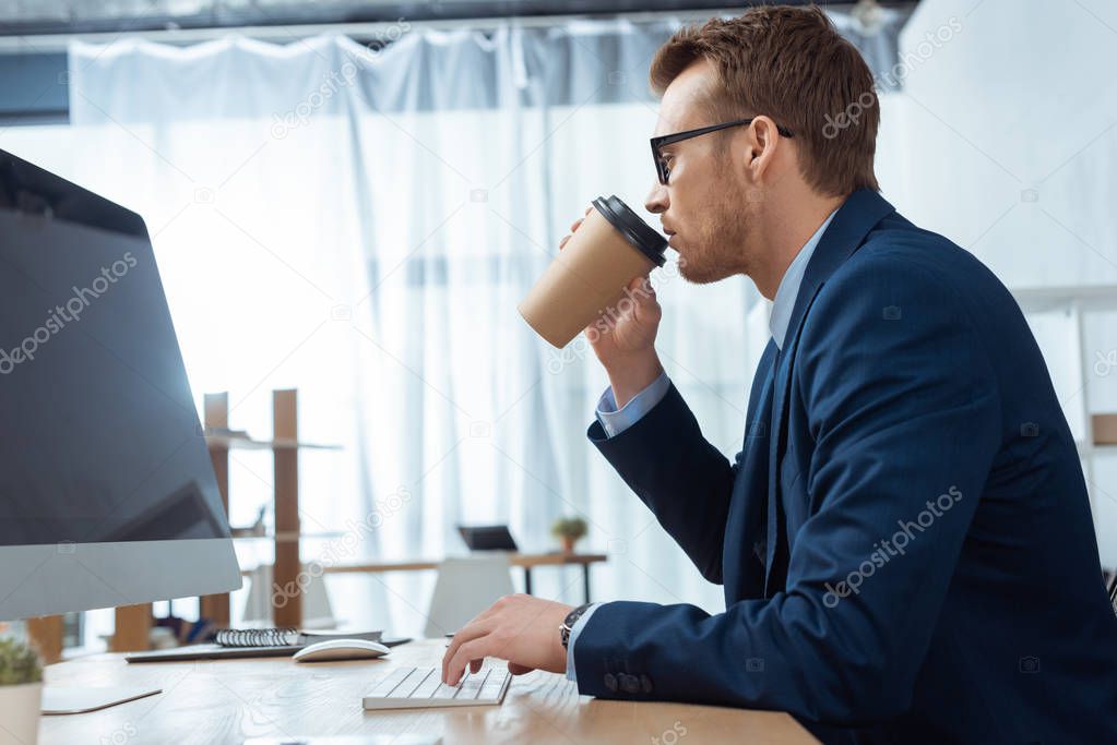 Side view of young businessman in eyeglasses drinking coffee and working at table with computer monitor in modern office