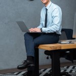 Laughing businessman sitting on table and working on laptop in modern office