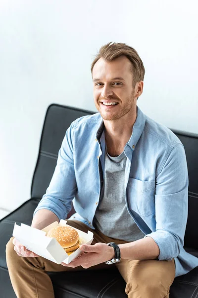 Sorridente Jovem Empresário Almoçando Com Hambúrguer Enquanto Sentado Sofá Escritório — Fotografia de Stock