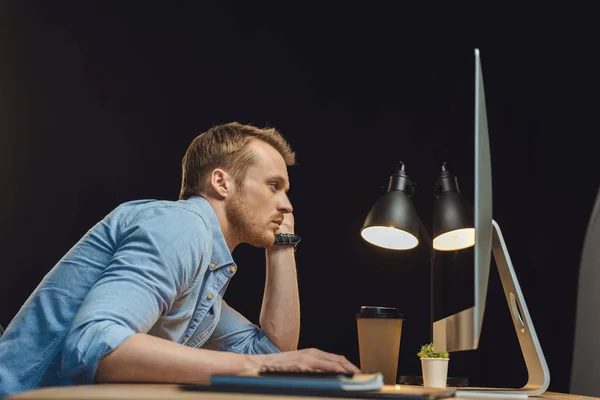 side view of overworked young businessman sitting at table with computer and disposable coffee cup under desk lump during late night in modern office
