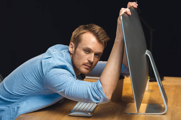 overworked young businessman holding computer monitor and looking at camera during night time in office 