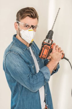 young man in protective goggles and mask holding electric drill and looking at camera clipart