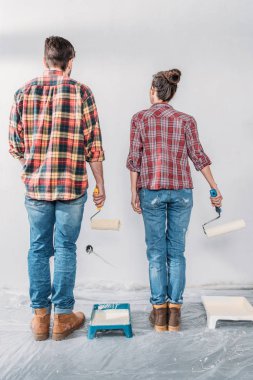 back view of young couple holding paint rollers and looking at wall in new house clipart