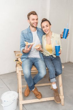 Kağıt bardak ve birlikte oturan ve yeni daire kamerada gülümseyen pizza dilimleri ile mutlu genç çift