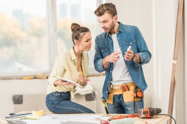 young couple using digital devices while making repairment in new apartment clipart