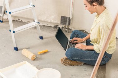 Yeni evde otururken dizüstü bilgisayar kullanan gülümseyen genç kadın görüntüsünü kırpılmış