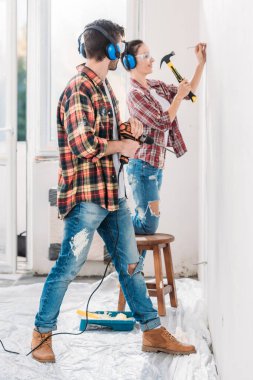 side view of young man holding electric drill and looking at smiling wife hammering nail in new apartment clipart