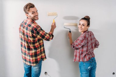 back view of happy young couple painting wall and smiling at camera clipart