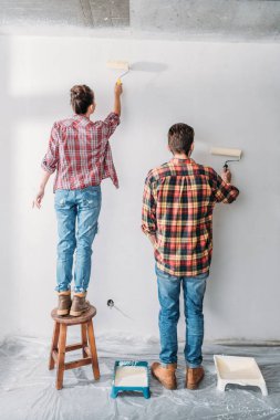 back view of young couple in checkered shirts painting wall in new apartment clipart