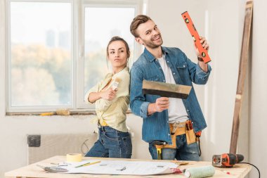 happy young couple holding renovation tools and smiling at camera in new apartment clipart