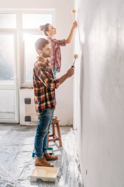 side view of young couple holding paint rollers and painting wall in new apartment
