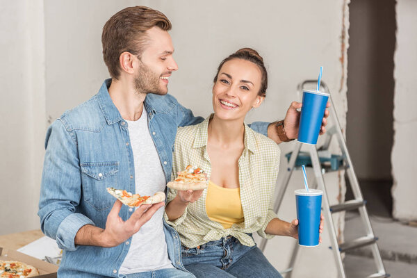 smiling young couple holding paper cups with drinking straws and pizza slices during repairment 