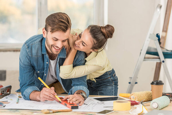 happy young woman hugging smiling boyfriend marking blueprint with level tool during house repair