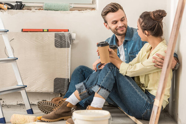 young couple holding paper cups and smiling each other while sitting on floor during house repair