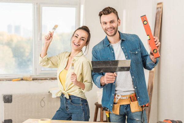 happy young couple holding tools and smiling at camera during house repair